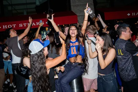 Getty Images A young woman wearing a Knicks jersey raises her hands in the air in celebration, surrounded by other young people.
