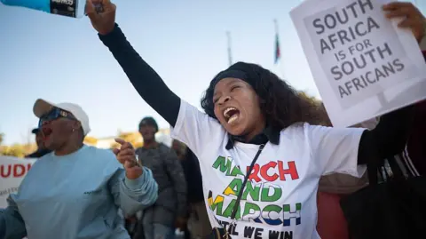 AFP via Getty Images A South African protester raises her fist during a demonstration; she is in a white T-shirt reading 'March and march until we win' and holding a placard with the words: 'South Africa is for South Africans'