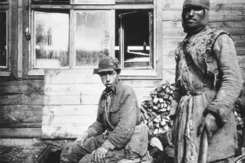 Alderney archives A black and white image on Alderney showing two prisoners of war wearing quilted cotton wool jackets and hats. The one in the centre is sitting, the one on the right standing and holding what looks like a farming tool. Behind them is a wooden hut with open windows.