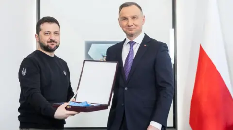 NurPhoto via Getty Images President Volodymyr Zelensky, bearded and wearing a black jumper, holds a box containing a medal next to then-President Andrzej Duda, who is wearing a suit and stands next to a red and white flag.
