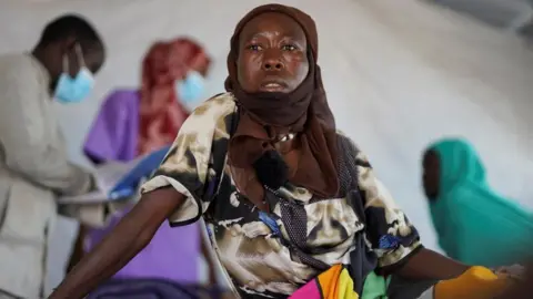 Reuters Halima Mohammed Adam, a Sudanese cholera patient with a brown shawl over her head and wearing a multi-coloured dress, looks on as she sits on a bed at a United Nations-run makeshift clinic in Tawila in Darfur, Sudan, on 5 August 2025.