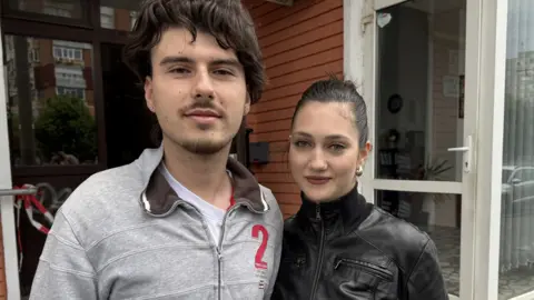 Adrian, wearing a white t-shirt and grey jacket, stands next to Ingrid, wearing a black leather jacket, outside the apartment block.