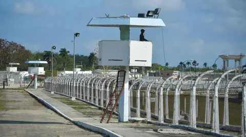AFP via Getty Images A prison tower overlooks lines of prison fencing. There is a blue sky in the background and a guard stands watch. 