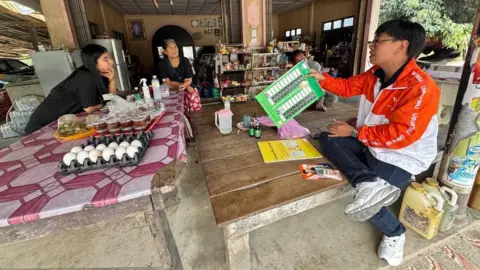 Chutiphong Pipoppinyo/Facebook Two women in a shop are talking to Chutiphong Pipoppinyo who is sitting on a bare wooden table and showing them a mockup of a ballot paper. Pipoppinyo is wearing blue jeans, white sneakers and an orange and white jacket. 