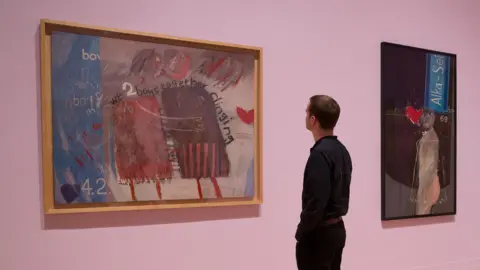 AFP via Getty Images An employee poses alongside artworks entitled 'We Two Boys Together Clinging, 1961' (L), 'The Most Beautiful Boy in the World, 1961' (C), and 'Tea Painting in an Illusionistic Style, 1961', by British artist David Hockney, during a photocall to promote a retrospective of Hockney's work, at the Tate Britain in London on February 6, 2017.