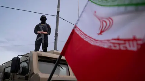 Getty Images An armed Iranian police officer stands guard on an armored vehicle, monitoring the area during a state-run religious rally in downtown Tehran, Iran, on April 29, 2026. An Iranian flag is seen in the foreground.