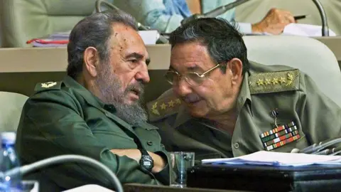 AFP via Getty Images Fidel Castro and his brother Raul are seen seated wearing military uniforms. Fidel is speaking and Raul is leaning in to hear him. 