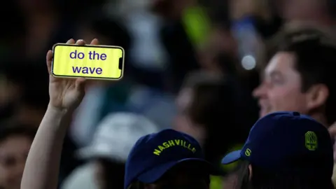 Getty Images A person whose face we cannot see wears a blue baseball cap and holds aloft a mobile phone with the words 'do the wave' on it. There is a blurry photo in the background of someone smiling. This appears to be a sports stadium full of people. 