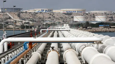 EPA View of Kharg Island oil terminal, showing a row of pipes stretching into the distance towards a raised area of ground where several silos sit, one of them has the words 