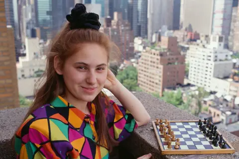 Sygma via Getty Images Judit Polgar as a teenager pictured on a balcony of a high rise building in a big city. She is standing next to a small chess board, wearing a colourful shirt. Her hair is in a high ponytail with a large scrunchie. She is smiling at the camera.