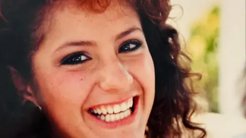 Lourdes Heredia Lourdes Heredia laughs and looks sideways to the camera. She has brown, curly hair and has a big smile.