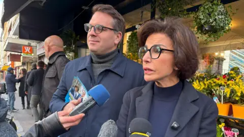 BBC/Paul Pradier A woman with black glasses speaks into microphones while a campaigner in a blue coat stands to her left