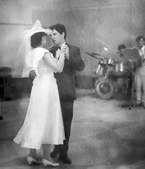 Supplied Iryna and Serhiy dance on their wedding day. She is wearing a white hat, a floaty white dress and white slip-on shoes with low heels. He is wearing a dark suit. He is looking into her eyes but with an anxious expression. 