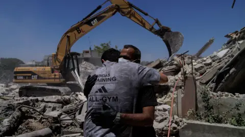 Reuters A man wearing a grey Adidas t-shirt hugging another man wearing a black t-shirt, gloves and surgical mask standing in front of a digger clearing up rubble in the aftermath of an Israeli airstrike in southern Lebanon.