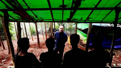 BBC/Darren Conway Four young men stand with their backs to the camera, in a jungle under a tarpaulin roof. BBC correspondent Quentin Sommerville stands facing towards them. 