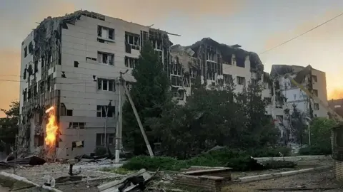 Reuters A large grey boxy building is visibly damaged and surrounded by debris and rubble. Flames are visible on one side of the building.