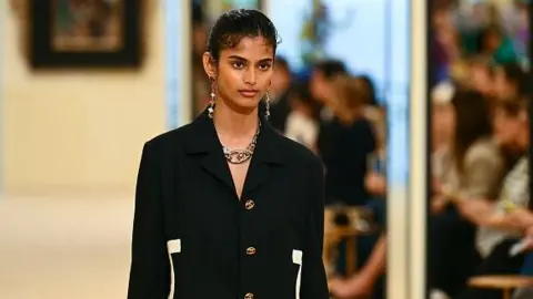 AFP via Getty Images Indian fashion model Bhavitha Mandava presents a creation for the Chanel Cruise 2026/2027 collection in Biarritz on April 28, 2026. She's wearing a black blazer with white detailing on the sides and golden buttons. 