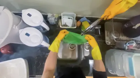 Shift Point of view from a camera on a person's head showing them wearing yellow rubber gloves and holding a green washcloth under the tap in a kitchen sink