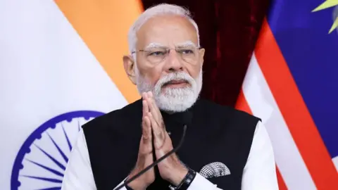 EPA Prime Minister Narendra Modi holds his hands together in a prayer style, as he stands before a microphone. Behind him can be seen the flags on India and Malaysia. 