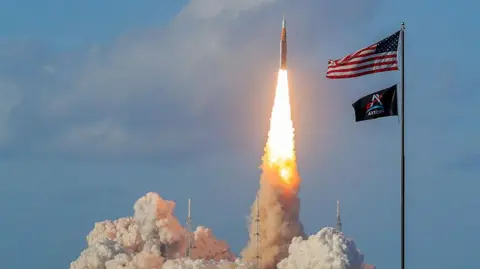 Getty Images NASA's Artemis II Space Launch System rocket and Orion spacecraft blasts off from the Kennedy Space Center. A vertical pillar of flame erupts from the rocket as it shoots off into the blue sky.
