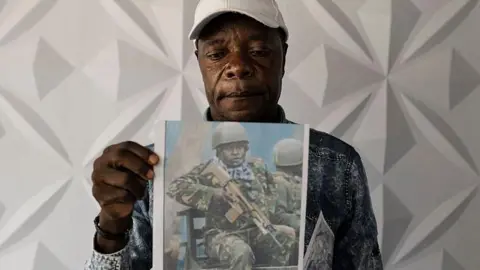 AFP via Getty Images Man wearing white baseball cap poses with a photo of man in army clothing holding a rifle.
