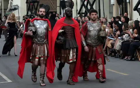 Getty Images Three male performers in red and brown gladiator costumes walk next to each other along the runway during Vogue World: Hollywood 