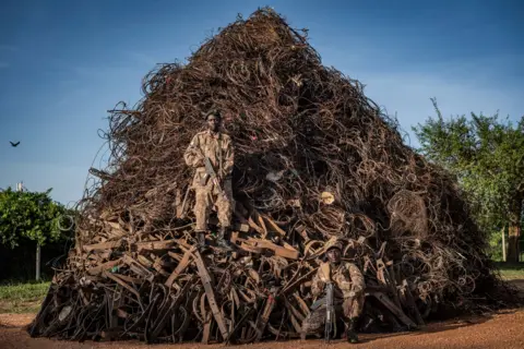 Adam Oswell / Wildlife Photographer of the Year A large mound of tangled metal wires, traps, and debris outdoors. One person stands on the wires in the middle and another knees beside the pile, giving a sense of scale to the chaotic mass.
