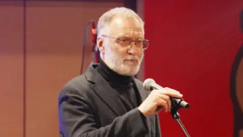 Russkaya Obshina Sergei Mikheev is wearing a grey suit and black polo neck and has glasses, short grey hair and a closely cropped grey beard. He is standing on a stage, holding a microphone.