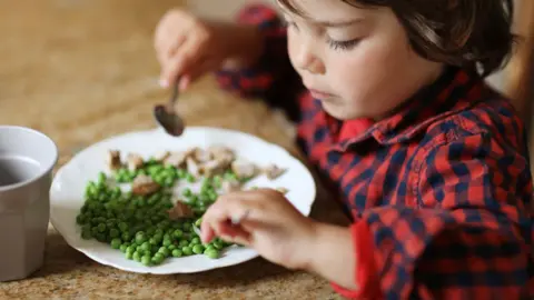 Getty Images A child with brown hair in a red and black chequered shirt sits at a table and eats a plate of peas and mushrooms