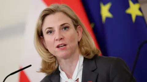 Reuters Headshot of Beate Meinl-Reisinger with shoulder-length blonde hair, black jacket and white shirt with pussy bow standing behind a microphone with an Austrian and EU flag in the background.