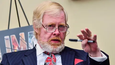 Getty Images A person in a dark pinstripe suit holding a pen, with a colourful paisley-patterned tie and a pocket square visible. 