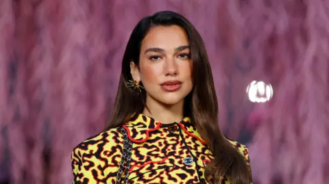 Reuters Dua Lipa poses with her dark brown hair straightened and out, while wearing a yellow, black and red Chanel jacket and a gold spiky earring. 