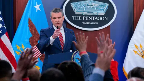 Getty Images Pete Hegseth calls on reporters during a press briefing at the Pentagon on 5 May. He is wearing a blue suit and red-striped tie and is holding a pen in his right hand as he points his index finger as reporters hold up their hands to ask a question
