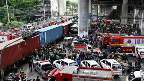 Getty Images A crowd gathers after a train crashed into a bus at a busy road crossing. There are several red firefighting trucks parked around the train.