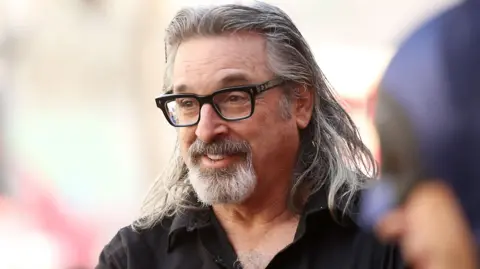 Getty Images Robert Carradine attends the ceremony honouring Burt Ward with A Star on the Hollywood Walk of Fame held on 9 Januray 2020 in Hollywood, California. He has shoulder-length grey hair and goatee beard and is wearing a black shirt and black glasses.