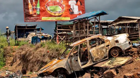AFP via Getty Images The wreckage of burnt-out car destroyed in an attack by suspected separatist fighters at the edge of the abandoned market in an Anglophone area of Cameroon. A soldier can be seen in the background.