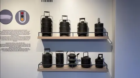 Shahid Sheikh Traditional metal lunchboxes displayed on two shelves in a museum, showing different shapes and sizes used in tiffin delivery systems.