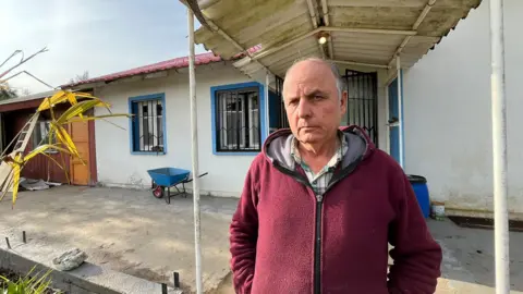Grace Livingstone Georg Klaube, in a red hoody, stands in front of a house in Villa Baviera. A blue wheelbarrow can be seen standing in front of a wall. The house is painted white and has blue accents around the windows. 