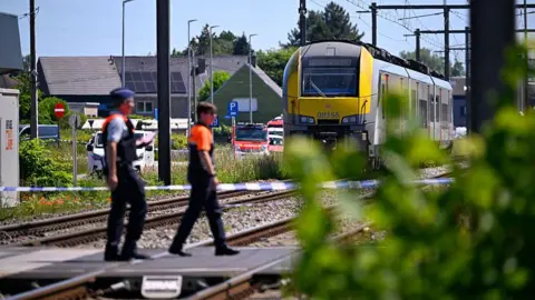 AFP via Getty Images olice officers walk at the site of an accident after a train (R) crashed into a school bus, at the railway crossing Vierhuizen in Buggenhout, some 20kms north of Brussels on May 26, 2026