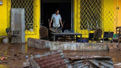EPA/Shutterstock A man stands in the doorway of a bright yellow building, in front of him the ground is completely covered in mud with twisted corrugated metal, broken furniture and scattered rubbish. Tables and chairs are also piled up and splattered in mud.