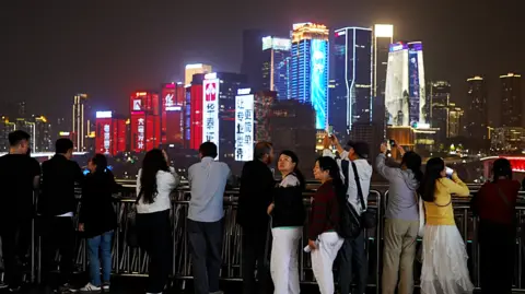 Male and female tourists take pictures of Chongqing's neon-lit skyscrapers, against a night-time sky.