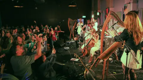 Connor Kenny Musicians perform on a small stage with violins, harp, guitar, and cello while a crowd stands closely in front, raising hands and recording on phones under warm stage lighting.
