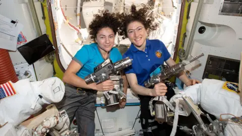 NASA Astronauts Jessica Meir and Christina Koch float side by side inside the space station, their hair fanned out in weightlessness. Jessica, on the left in a turquoise T‑shirt, and Christina, on the right in a blue polo shirt, each hold a large silver power tool used to service spacesuits. Around them are white spacesuit parts, cables and panels, giving a busy workshop feel inside the curved, pale interior of the airlock module.