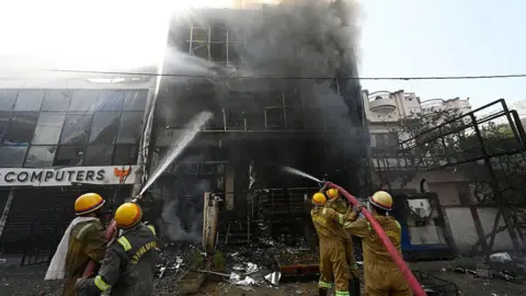 Getty Images Five firefighters spray water on the burnt building in India's Lucknow. Thick, dark smoke can be seen billowing from the structure.