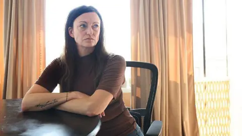 Toronto Star via Getty Images Claire Brosseau is photographed at her home in Toronto, Canada. She has dark brown hair and blue eyes. She is seated at a table with her arms crossed. She is wearing a brown short-sleeved turtle neck.