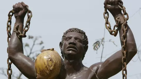 AFP via Getty Images Close up of statue showing liberation from slavery on Goree Island in Senegal. It is a man from shoulders up, with his arms raised, holding a broken chain in his hands which is still attached to his wrists. The statue also includes the figure of a woman standing in front of the man - all that is visible of her is the back of her head, and she has a head wrap on.