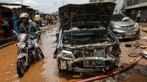 EPA A man rides a motorbike through brown flood water next to a damaged car after it was swept away by floods at Grogon garage area, known for automotive workshops and auto spares along the banks of the Nairobi River in downtown Nairobi, Kenya, 07 March 2026.