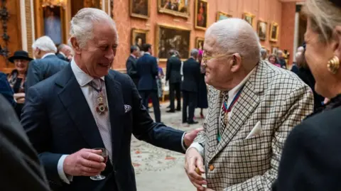 Getty Images King Charles III talks with Mr David Hockney during a luncheon for Members of the Order of Merit at Buckingham Palace on November 24, 2022 in London, England. The King has his hand on the arm of Hockney. David Hockney is wearing a distinctive suit with the two men smiling at each other. 