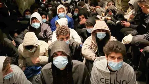 Kurdish Regional Government A large group of young men wearing hooded sweatshirts and light-blue surgical masks sit closely together on the floor in a crowded indoor space. Most face forward toward the camera, with some looking downward or to the side. The setting appears dimly lit, with people packed tightly in rows, filling the frame from foreground to background. Clothing is mostly dark or neutral tones, and the confined arrangement suggests limited space.