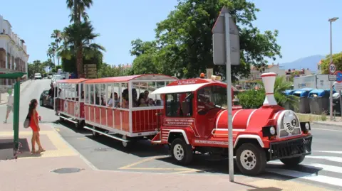 Ayuntamiento Cártama A red tourist train driving down a sunny street, with several people onboard